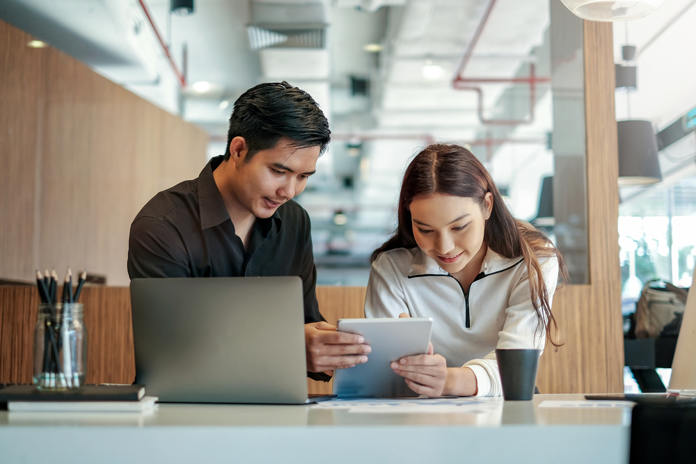 Um homem, e uma homem, um notbook na mesa, e a mulher segurando um tablet